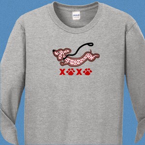 May include: A gray long-sleeved t-shirt with a cartoon dachshund wearing a red heart pattern and a leash. The dachshund is running and the text "XOX" is below the dog with paw prints.