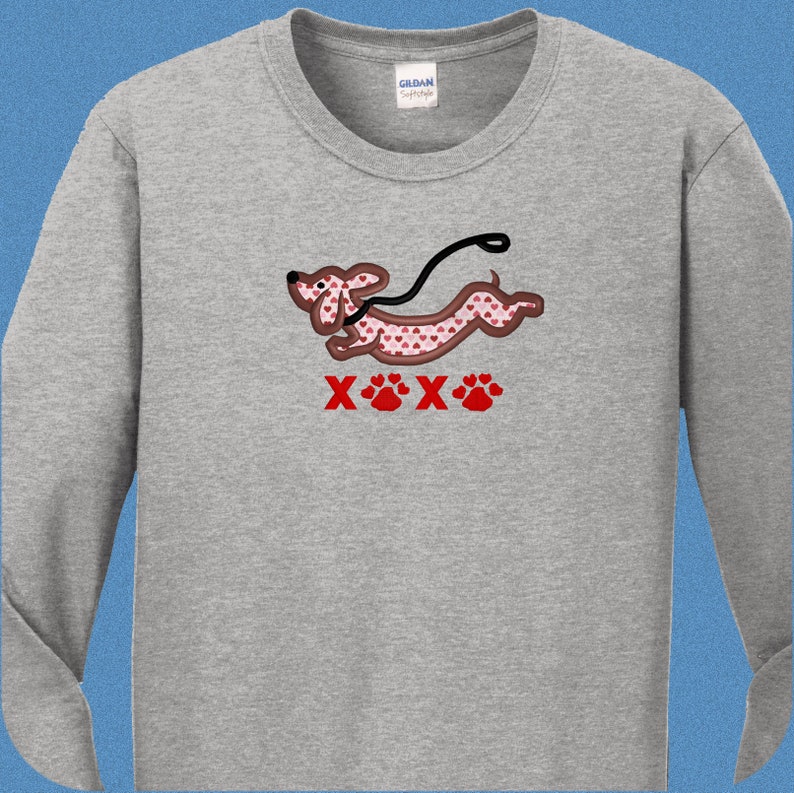 May include: A gray long-sleeved t-shirt with a cartoon dachshund wearing a red heart pattern and a leash. The dachshund is running and the text "XOX" is below the dog with paw prints.