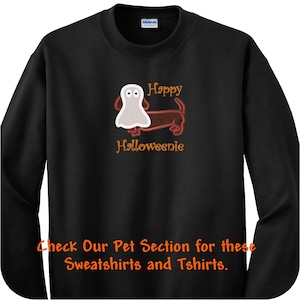 May include: A black long-sleeve sweatshirt featuring a brown dachshund dog wearing a white ghost costume. The text above the dog reads Happy and below the dog reads Halloweenie in orange. The bottom text reads Check Our Pet Section for these Sweatshirts and Tshirts.