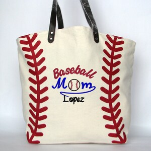 A large off white canvas tote bag with printed red baseball stitching on the sides. The bag has black faux leather handles attached with silver rivets. The front of the bag has a three line monogram:  top line is 'BASEBALL' in red thread, middle line is 'MOM' in blue thread and bottom line is  'Lopez' in blue thread and all text is embroidered in script font.  An embroidered baseball is replacing the 'O' in   'MOM' using the Script Mom Name design.