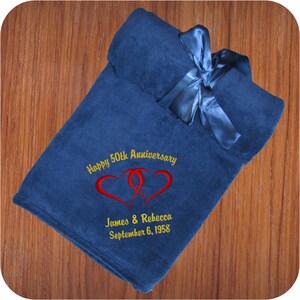 A soft blue fleece throw blanket with a satin ribbon tied around it. The ribbon has a hook & loop closure for easy gifting and/or quick storage. This 50th anniversary throw blanket has two embroidered red intertwined hearts with 'Happy 50th Anniversary' above and 'James & Rebecca September 6, 1958' in 2 line below all in gold using the Anniversary Hearts design.  The custom monogram is located in the bottom corner of this fleece throw and makes an elegant and cozy anniversary or wedding gift.