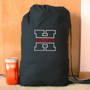May include: A black drawstring laundry bag with the name "HARRIS" embroidered in red within a white outlined letter "H". An orange container with a white lid sits to the left of the bag. The bag is made of a dark fabric.