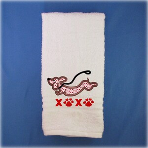 May include: White hand towel with an embroidered image of a dachshund running with a leash. The dachshund is pink with a brown outline and has a pattern of small pink hearts. The text "XXX" is embroidered in red below the dachshund, with three red paw prints.