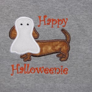 May include: A heather gray t-shirt featuring an embroidered brown dachshund dog wearing a white ghost costume over its head. The text "Happy" is embroidered in orange above the dog, and the word "Halloweenie" is embroidered in orange below.