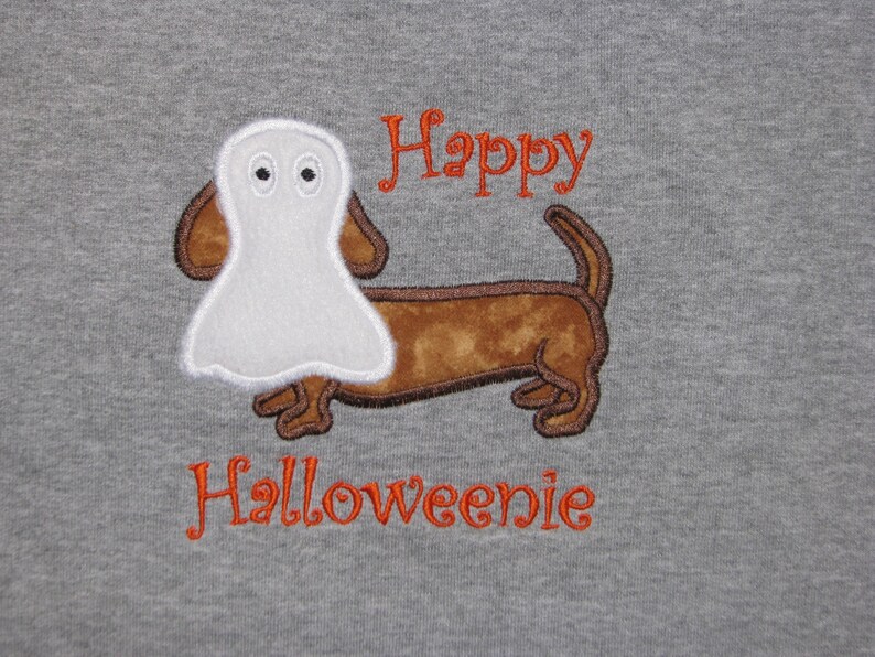 May include: A heather gray t-shirt featuring an embroidered brown dachshund dog wearing a white ghost costume over its head. The text "Happy" is embroidered in orange above the dog, and the word "Halloweenie" is embroidered in orange below.