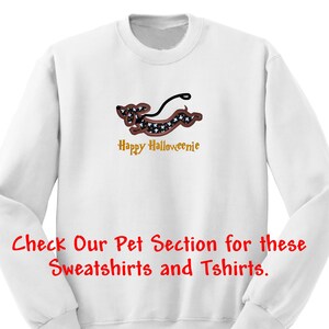 May include: A white long-sleeve sweatshirt featuring an embroidered dachshund dog with a black and white patterned body, a brown outline, and a black leash. Below the dog, the text reads Happy Halloweenie in gold. Red text at the bottom reads Check Our Pet Section for these Sweatshirts and Tshirts.