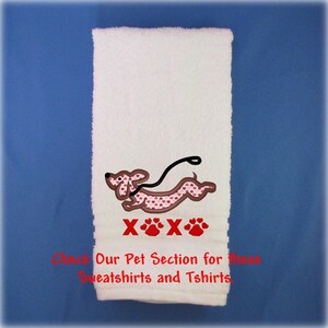 May include: A white hand towel featuring an embroidered brown dachshund with a pink heart pattern, a black leash, and red text reading XOXO with paw prints. Below, red text reads: Check Our Pet Section for these Sweatshirts and Tshirts.
