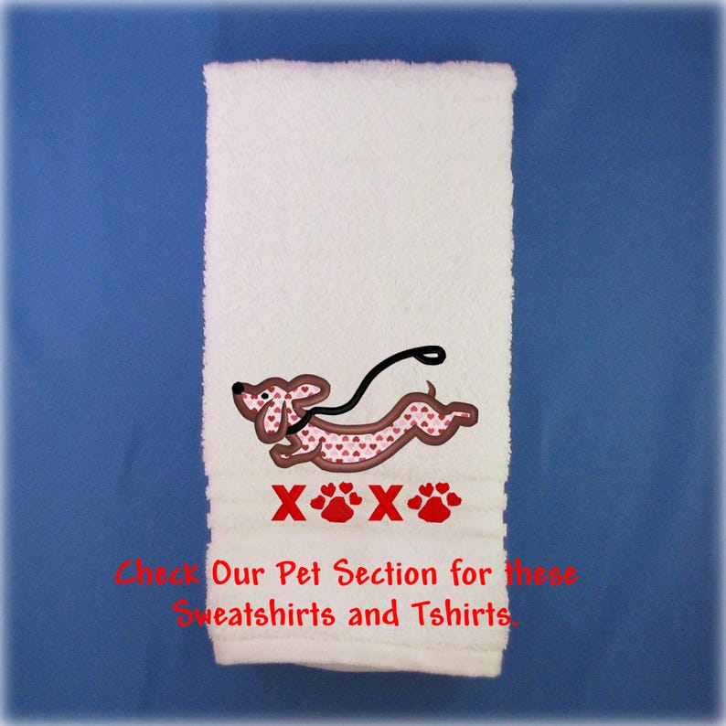 May include: A white hand towel featuring an embroidered brown dachshund with a pink heart pattern, a black leash, and red text reading XOXO with paw prints. Below, red text reads: Check Our Pet Section for these Sweatshirts and Tshirts.