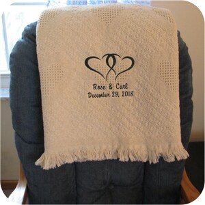 A natural colored personalized throw blanket folded in thirds and over a chair to show how big it is.  Two intertwined dark green hearts are embroidered above 'Rose & Carl' and 'December 29, 2018' in 2 lines all in dark green thread using the Double Hearts design.  The monogram is embroidered on the bottom center of a heart weave cotton throw and makes the perfect elegant wedding or anniversary gift.