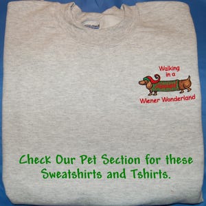 May include: A light gray crewneck sweatshirt featuring an embroidered dachshund wearing a red and green elf hat and a green sweater. The text reads Walking in a Wiener Wonderland and Check Our Pet Section for these Sweatshirts and Tshirts.