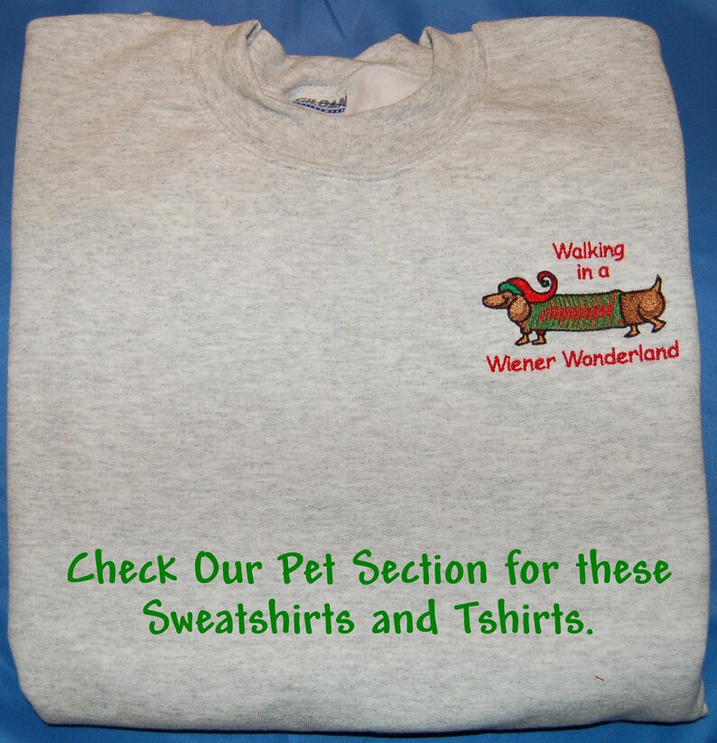 May include: A light gray crewneck sweatshirt featuring an embroidered dachshund wearing a red and green elf hat and a green sweater. The text reads Walking in a Wiener Wonderland and Check Our Pet Section for these Sweatshirts and Tshirts.