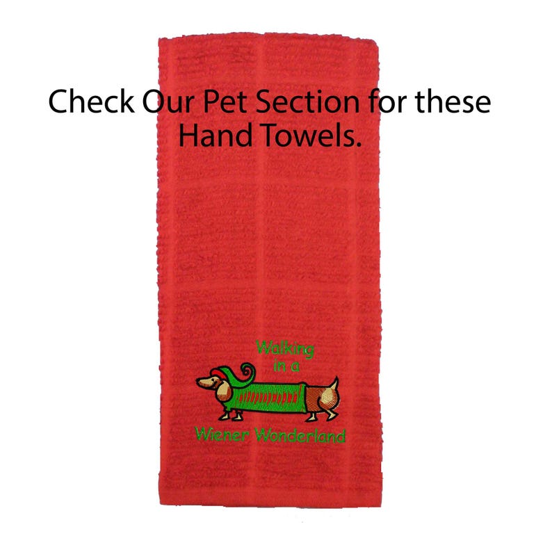 May include: A red hand towel featuring an embroidered dachshund wearing a green sweater and a red hat. The text reads "Walking in a Wiener Wonderland" and "Check Our Pet Section for these Hand Towels." The towel has a textured, grid-like fabric pattern.