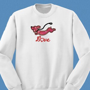 May include: White sweatshirt with a red dachshund with a heart pattern and a red leash. The dachshund is running and the text "Love" is written below it in red.