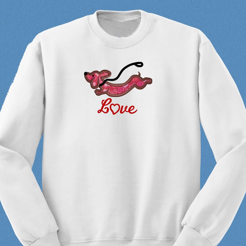 May include: White sweatshirt with a red dachshund with a heart pattern and a red leash. The dachshund is running and the text "Love" is written below it in red.