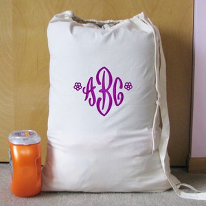 An white canvas drawstring bag with a purple thread monogram 'ABC' and small flowers on both ends embroidered with End Scroll Flower design. The bag is sitting in front of a closet on a light brown carpet next to a container of laundry detergent.