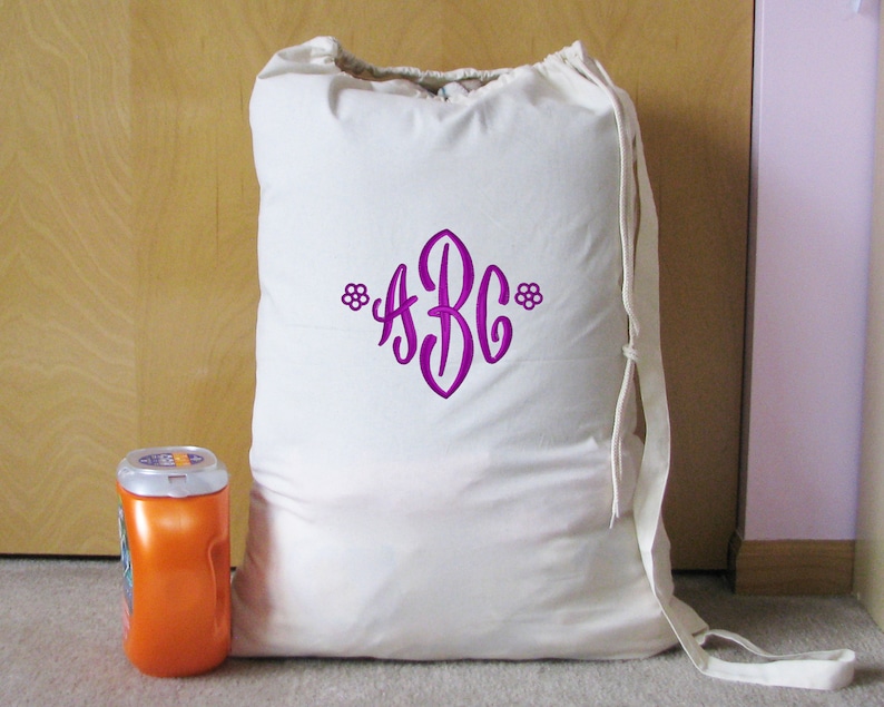 An white canvas drawstring bag with a purple thread monogram 'ABC' and small flowers on both ends embroidered with End Scroll Flower design. The bag is sitting in front of a closet on a light brown carpet next to a container of laundry detergent.