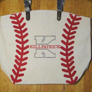 A large off white white canvas tote bag with printed red baseball stitching and a gray embroidered large letter 'K' with the name 'KILLPATRICK' in red thread in the middle of the 'K' using the Split Letter design.  The bag also has black faux leather straps.