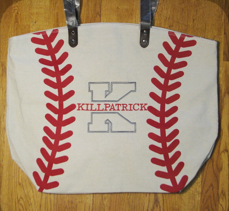 A large off white white canvas tote bag with printed red baseball stitching and a gray embroidered large letter 'K' with the name 'KILLPATRICK' in red thread in the middle of the 'K' using the Split Letter design.  The bag also has black faux leather straps.