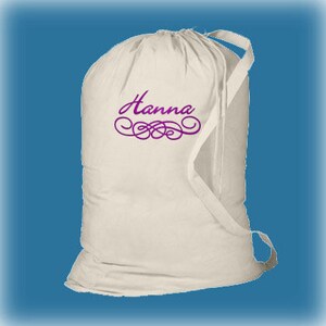 An off white canvas laundry bag with shoulder strap with a purple embroidered 'Hanna' and a purple swirl below using the Swirl border design.