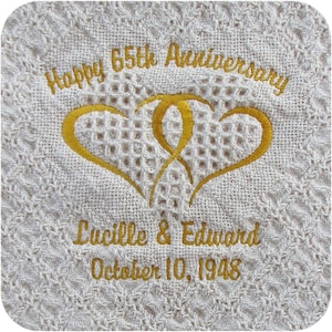 A closeup of a natural colored personalized throw blanket with two embroidered gold intertwined hearts with 'Happy 65th Anniversary' above and 'Lucille & Edward' and 'October 10, 1948'  in 2 lines below all in gold thread using the Anniversary Hearts design.  The monogram is embroidered on the bottom center of a heart weave cotton throw.