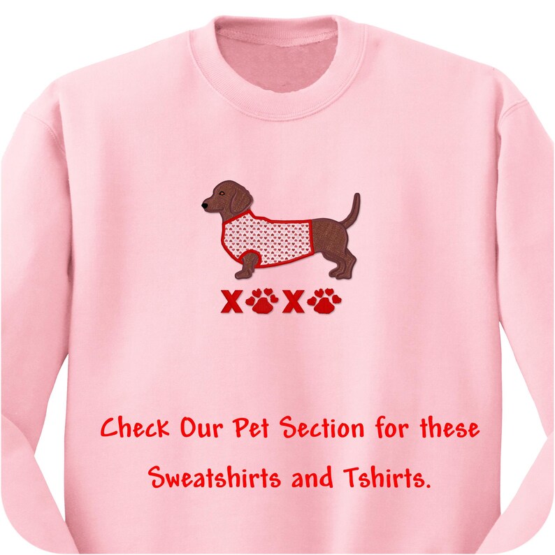 May include: A light pink crewneck sweatshirt featuring an embroidered brown dachshund wearing a white sweater with red hearts. Below the dog are red XOXO letters and paw prints. The text reads: Check Our Pet Section for these Sweatshirts and Tshirts.