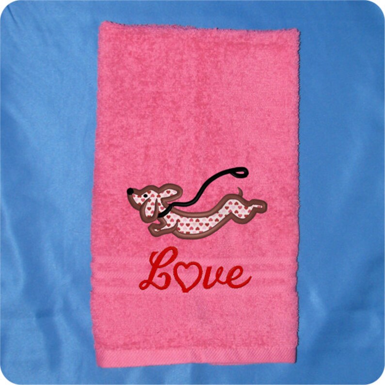 May include: Pink towel with an embroidered dachshund wearing a heart-patterned shirt and a leash. The word "LOVE" is embroidered below the dog with a heart in the "O".