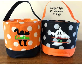 May include: Two cylindrical fabric buckets with handles. The left bucket is orange with white polka dots, a black base, and a black cat silhouette with the name Caroline. The right bucket is black with white polka dots, an orange base, and a white ghost with the name Ty, labeled Large Style 10" diameter 9" high.
