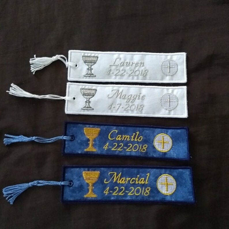 May include: Embroidered bookmarks in white and blue with tassels. The white bookmarks have silver chalice and cross designs, with names and dates. The blue bookmarks feature gold chalices, names, dates, and crosses.
