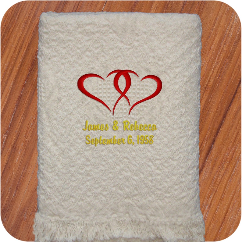 A natural colored personalized throw blanket with two embroidered gold intertwined hearts with 'James & Rebecca' and 'September 6, 1958' in 2 lines below all in gold using the Double Hearts design.  The monogram is embroidered on the bottom center of a heart weave cotton throw and makes the perfect elegant wedding gift.