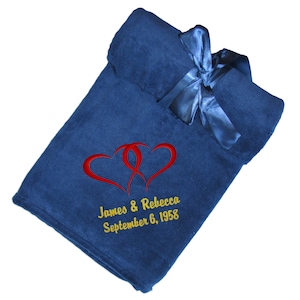A super soft blue fleece throw blanket with a satin ribbon tied around it. The ribbon has a hook & loop closure for easy gifting and/or quick storage. The wedding or anniversary fleece blanket has two embroidered red intertwined hearts with 'Happy 50th Anniversary' above and 'James & Rebecca September 6, 1958' in 2 line below all in gold using the Double Hearts design.  The monogram is located in the bottom corner of this fleece throw and makes an elegant and cozy anniversary or wedding gift.