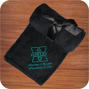 A black embroidered monogram fleece blanket that comes with a matching decorative satin ribbon with hook & loop closure for easy gifting and/or quick storage.  A large outline 'H' is embroidered with the name 'HAWLEY' located in the middle of the H using the Split Letter design.  Below the monogram in 2 lines is 'Andrew & Karen' and 'November 19, 2016'.  The custom monogram is located in the bottom corner of this fleece throw and makes the perfect elegant and cozy wedding or anniversary gift.