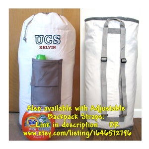 An extra large, heavy duty white laundry bag with gray trim and gray zippered pocket containing laundry detergent is in front of a closet. The back of the bag has adjustable backpack straps and carrying handle.  Varsity Initials are in navy and red thread.