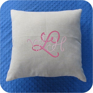 Puede incluir: Una almohada de lino beige con un monograma bordado en rosa de las letras "LH" en una fuente cursiva.