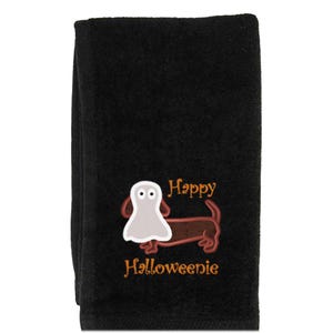 May include: A black hand towel featuring an embroidered brown dachshund dog wearing a white ghost costume over its head. The text "Happy Halloweenie" is embroidered in orange lettering above and below the dog graphic on the towel fabric.