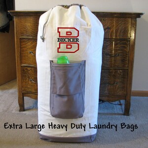 An extra large heavy duty white laundry bag with gray trim and gray zippered pocket containing laundry detergent is in front of a bedroom dresser. The laundry bag is embroidered with the Split Letter design, a large letter B in red thread and recipient's name "BECKER" in the middle with black thread.
