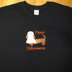 May include: A black short-sleeved t-shirt featuring an embroidered brown dachshund dog wearing a white ghost costume. The text "Happy" is embroidered in orange above the dog, and the text "Halloweenie" is embroidered in orange below the dog.
