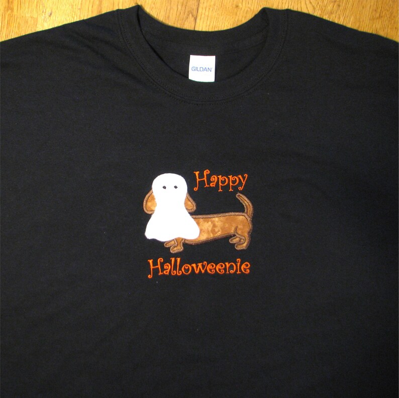 May include: A black short-sleeved t-shirt featuring an embroidered brown dachshund dog wearing a white ghost costume. The text "Happy" is embroidered in orange above the dog, and the text "Halloweenie" is embroidered in orange below the dog.