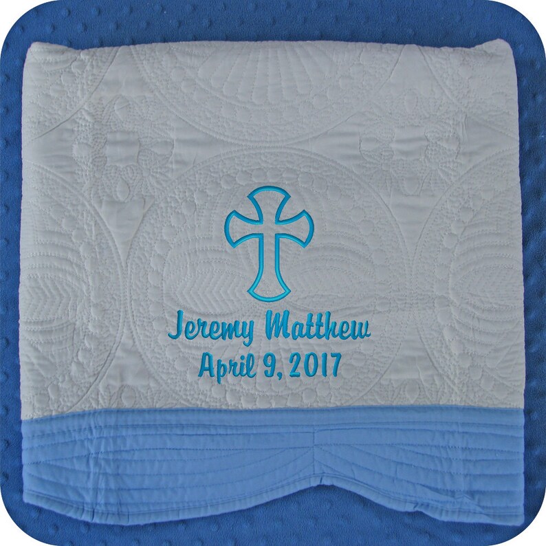 Pode incluir: Cobertor de beb&ecirc; acolchoado branco com borda azul. O cobertor tem uma cruz bordada em azul e o texto "Jeremy Matthew April 9, 2017".