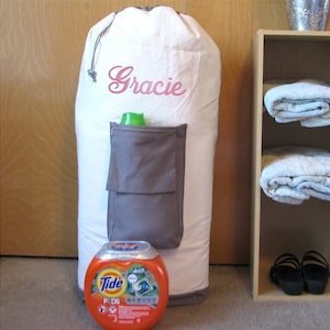 A extra large, heavy duty white laundry bag with gray trim and gray zippered pocket containing laundry detergent is in front of a closet. The laundry bag is personalized with "Gracie" in the swirly Diane script font in pink thread.