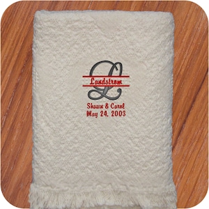 Natural colored cotton heart weave patterned throw blanket with fringe. A custom embroidery design on the blanket is a large split script L in dark gray thread with couples last name 'Landstrom' in red thread in the middle.  The couples first names and wedding date in red thread below the L.  The personalized design in on the bottom center of the wedding throw and makes an elegant wedding gift.