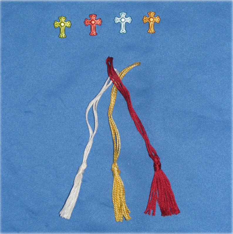 May include: Four decorative cross charms in yellow, red, light blue, and gold are displayed above four braided tassels in white, gold, and red. The tassels are arranged on a blue background, creating a visually appealing arrangement.