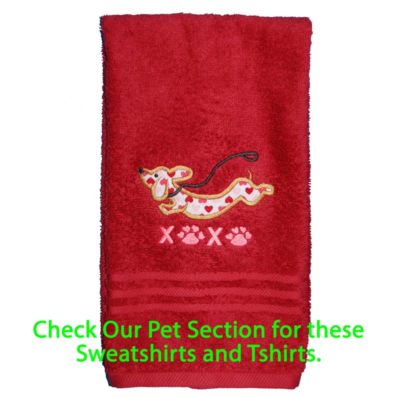 May include: A red hand towel featuring an embroidered dachshund dog with a white body covered in small red and pink hearts. Below the dog, the text reads X paw print X paw print. The green text at the bottom says Check Our Pet Section for these Sweatshirts and Tshirts.
