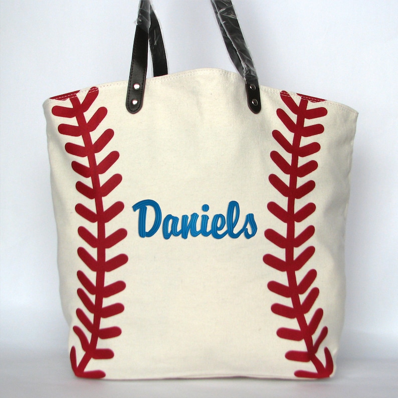 A large off white canvas tote bag with printed red baseball stitching on the sides. The bag has black faux leather handles and a blue embroidered name 'Daniels' in Brody script font.