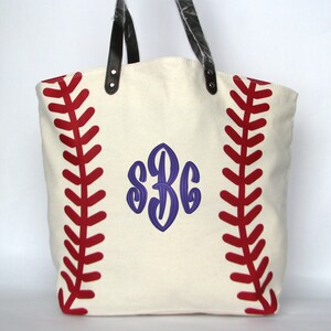 A large off white canvas tote bag with printed red baseball stitching on both sides. The bag is embroidered with a dark blue monogram 'S B C' in the center using the End Scroll NO Flower design.