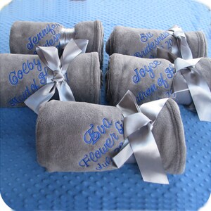 5 super soft gray fleece blankets embroidered with girls' names and their wedding role (Maid of Honor, Bridesmaid, Flower Girl) in a swirly script font and blue thread.  These elegant and cozy fleece blankets tied with a matching satin bow make the perfect wedding party gifts.
