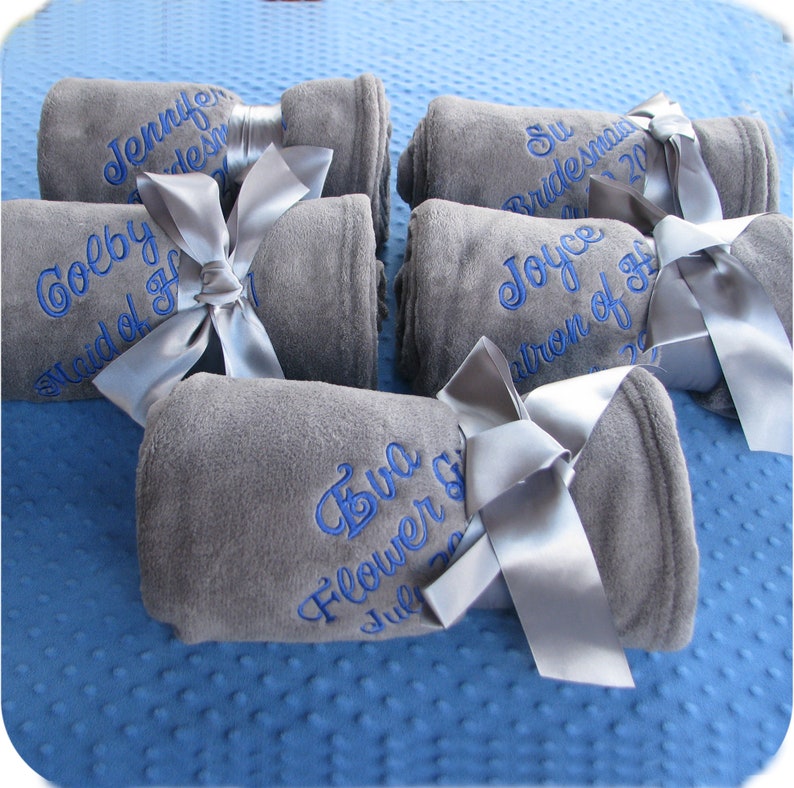 5 super soft gray fleece blankets embroidered with girls' names and their wedding role (Maid of Honor, Bridesmaid, Flower Girl) in a swirly script font and blue thread.  These elegant and cozy fleece blankets tied with a matching satin bow make the perfect wedding party gifts.