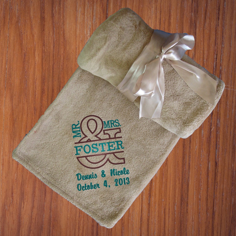A gray personalized fleece blanket that comes with a matching decorative satin ribbon. A large ampersand is embroidered in dark brown thread with the name 'FOSTER' in teal thread located in the middle using the Split & design.  'MR' is at the top left and 'MRS' at the top right of the ampersand and both in teal thread .  Below the monogram in 2 lines is 'Dennis & Nicole' and 'October 4, 2013' all in teal thread.  The custom monogram is located in the bottom corner of this fleece throw.