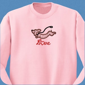 May include: Pink sweatshirt with a red dachshund with a heart pattern running with a leash. The text "Love" is below the dachshund.