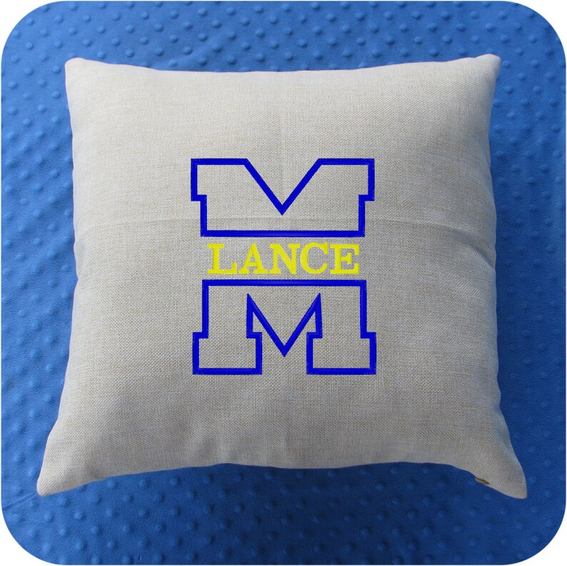 Puede incluir: Una almohada beige con una letra "M" bordada en azul y amarillo y la palabra "LANCE" en amarillo.