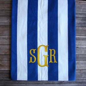 May include: Blue and white striped beach towel with a gold monogram. The towel features bold, vertical stripes and a three-letter monogram in a classic, elegant font. The towel is made of a soft, absorbent material.